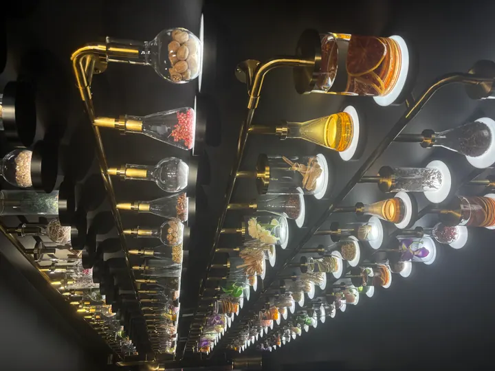 A wall of lit laboratory flasks filled with spices, flowers and wood.